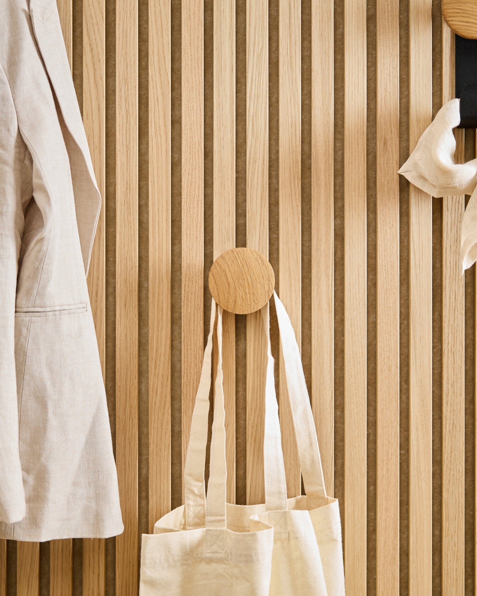 En beige rock och tygväska hänger på Linn I Single Hanger i Natural Oak från WoodUpp, monterad på en vertical Akupanel akustikpanel i trälamell.